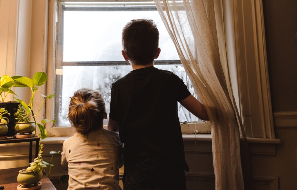 Two young children look out a living room window at a snow storm