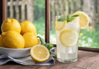 A bowl of lemons sits on a table next to a sliced lemon and a glass of lemonade.
