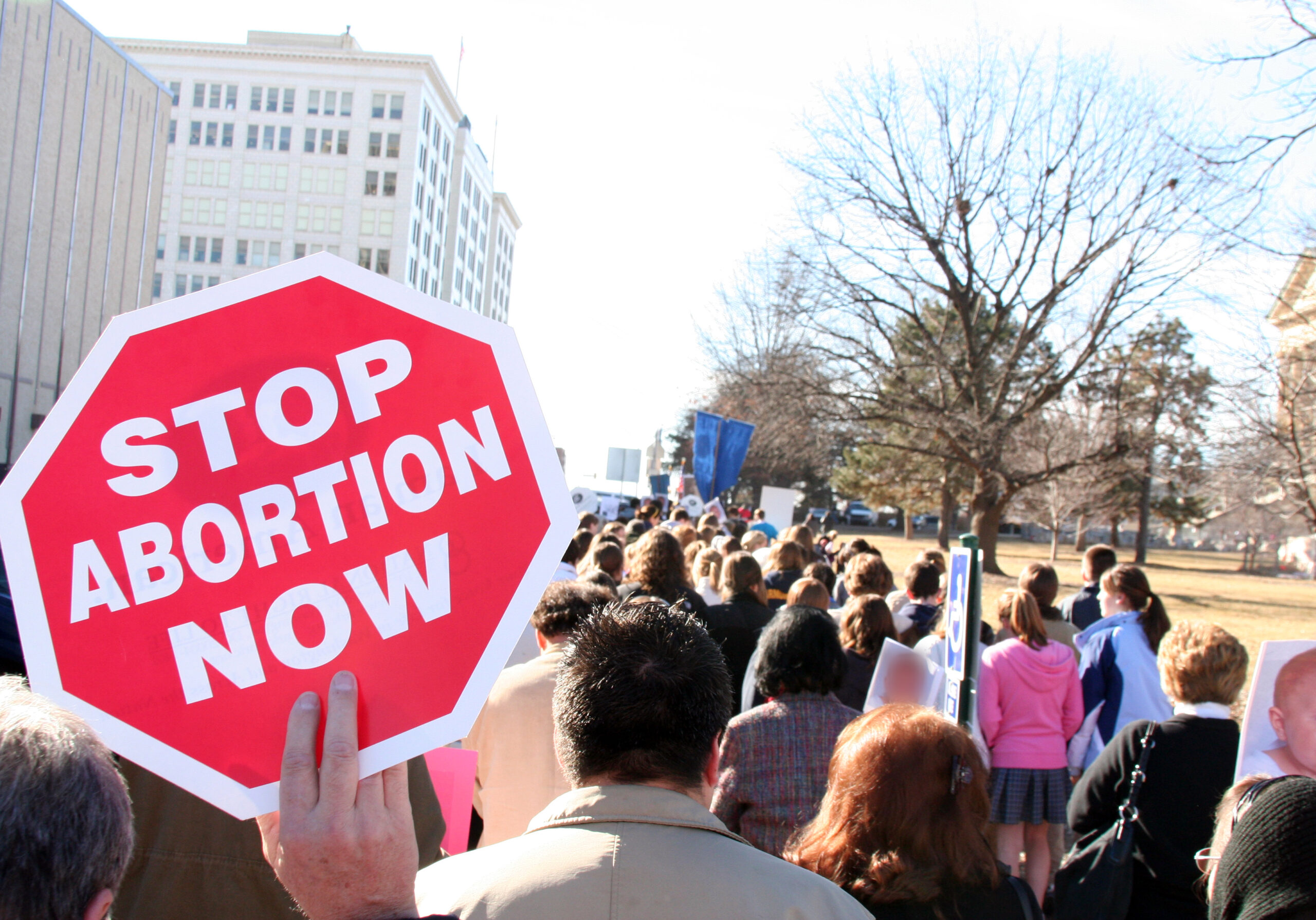 A group of people march on a sidewalk with one person holding a sign shaped like a STOP sign which reads "Stop Abortion Now."
