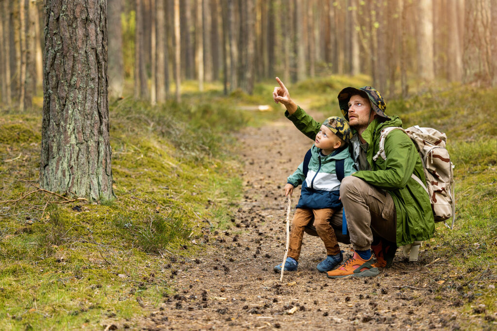 A father and son crouched in the woods during a hike. The father is pointing at something above.