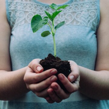 A white woman in a blue tank top is holding a handful of dirt. A plant is growing out of the dirt in her hands.