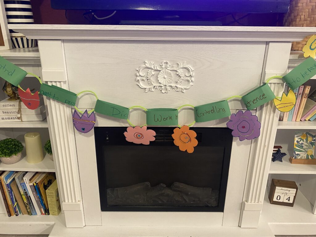 Ordinary Time garland with green paper and multicolored flowers hung on a fireplace between two white bookshelves. 