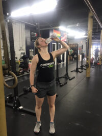 Woman drinking from a water bottle in the gym. She is wearing a CrossFit tank top.