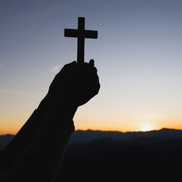 Someone holding a cross with both hands. The background features a sun setting over the mountains.