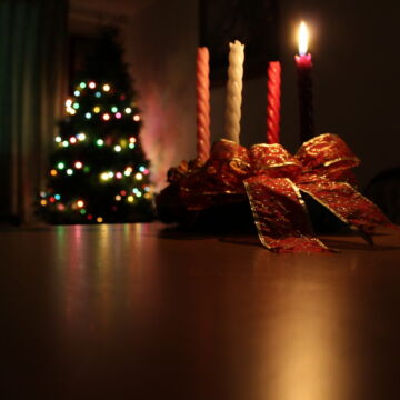 Dark room with Advent wreath on a tabletop with one candle lit and a blurry Christmas tree with lights on in background