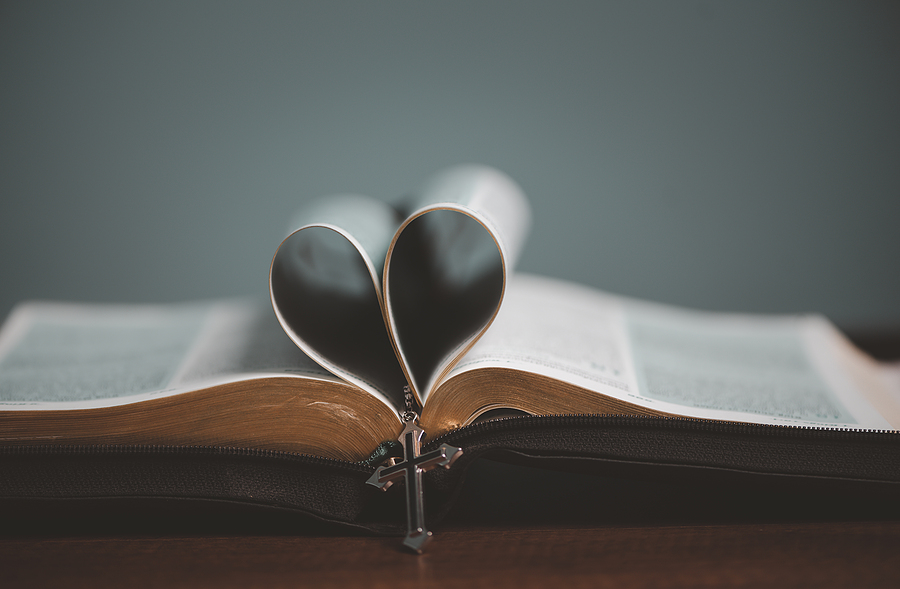Holy Bible open with pages folded in the middle to make a heart shape and cross charm haning from the heart.