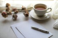 Paper and pen set out on white table. Behind it is a few light pink flowers and a white cup of tea. At the back of the image is a white sheet or curtain draped upon a table.